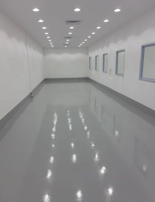 غرفة واسعة بأرضية إيبوكسي رمادية لامعة بالمدينة المنورة وجدران بيضاء مزودة بإضاءة سقفية حديثة، تُظهر لمسة احترافية في تنفيذ دهانات الإيبوكسي المناسبة للمصانع والمعامل والمستودعات