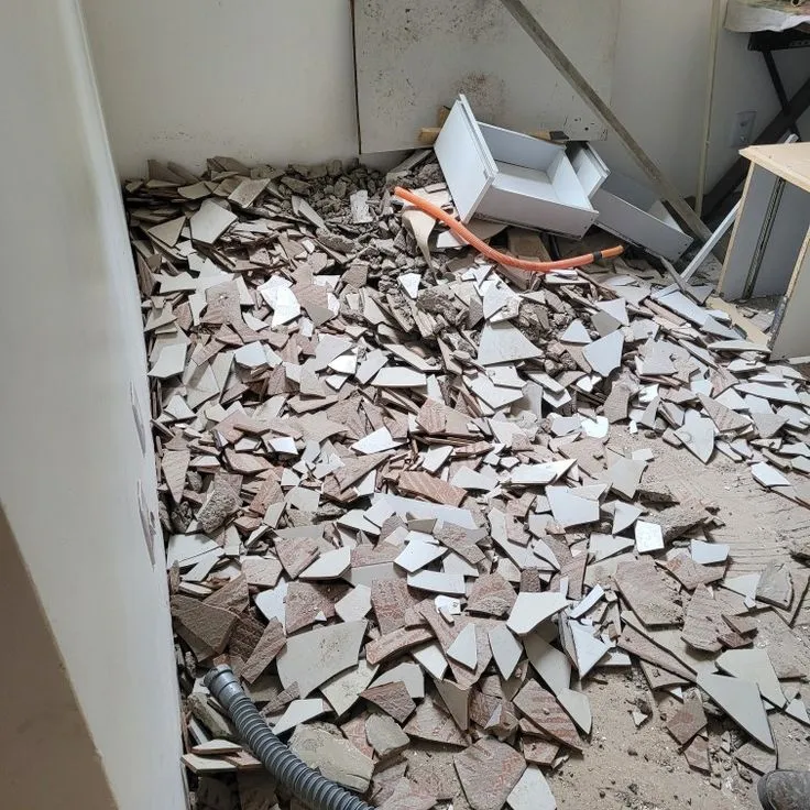 النص البديل المناسب لهذه الصورة هو: **أرضية مغطاة ببقايا سيراميك مكسور أثناء عملية إزالة البلاط ضمن أعمال ترميم منزل