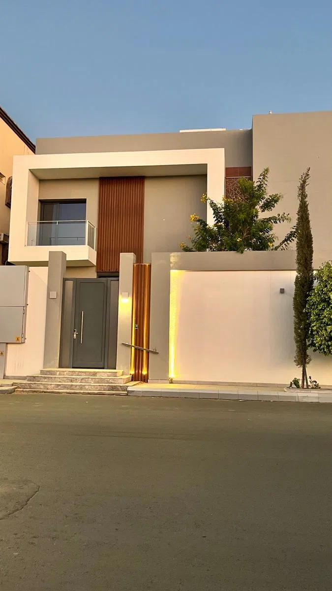 دهان واجهة منزل بدرجات البيج والرمادي مع لمسات خشبية وباب خارجي وإضاءة جدارية مخفية وأشجار زينة بجانب السور